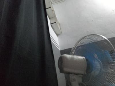 A close-up, low-angle, medium shot shows a black curtain on the left side of the frame, and a fan on the right. The fan has a silver grill and a blue motor housing. The wall behind the fan is white and textured. The scene appears to be indoors. The location in Senti, Nigeria, cannot be determined from the image.