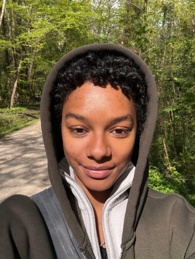 A person with dark hair and brown eyes, wearing a dark green hooded jacket over a light gray zip-up sweatshirt, is facing the camera in an outdoor setting. The person's face is well-lit, and they appear to be smiling slightly. The background consists of green trees and foliage, and a sunlit path is visible. Although there is no specific evidence indicating this image was taken in Noisiel, France, the natural landscape suggests a park or wooded area.