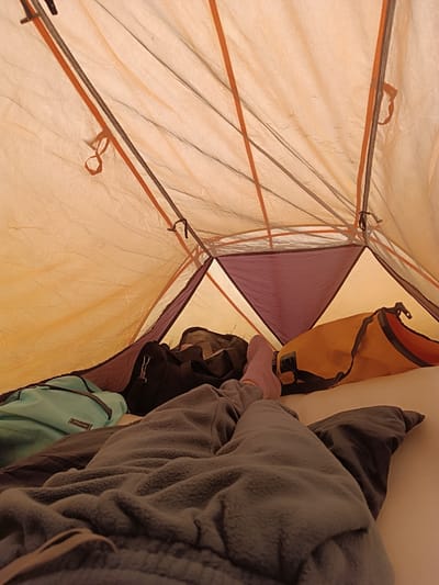 Inside a tent, a person's legs covered by a grey blanket are visible, extending towards the foreground. The person is wearing pink socks. The tent interior shows an orange ceiling with orange and brown poles, and segments of purple fabric. Bags are placed around the tent: a teal bag on the left, a black duffel bag behind it, and an orange bag on the right. The floor of the tent is covered with a white sheet. The scene is forensic and matter-of-fact. The location is Idrisovo, Russia.