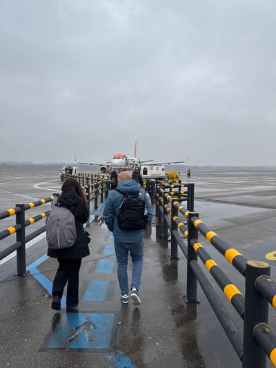 The image depicts an airport tarmac scene on an overcast, wet day at Milan Malpensa Airport in Somma Lombardo, Italy. In the foreground, two individuals walk away from the camera along a designated pedestrian pathway marked with blue symbols of a walking person and arrows. The path is bordered by black vertical posts with two horizontal bars featuring alternating black and yellow stripes.
The person on the left wears a dark, possibly quilted jacket, dark trousers, and a light grey backpack. The person on the right is male, bald, wearing a blue quilted jacket, blue jeans, and dark athletic shoes, carrying a black quilted backpack with light blue straps.
In the mid-ground, an EasyJet aircraft, identifiable by its white fuselage and orange tail with the airline's logo, is parked on the tarmac. A mobile passenger stair is connected to the aircraft's front left door. Various ground support equipment surrounds the aircraft, including a pushback tug near the nose and other service vehicles, one with a yellow hose or cable, towards the right. Several indistinct figures, some appearing to be airport personnel in high-visibility vests, are present around the aircraft. The tarmac is reflective due to moisture, indicating rain. The background shows a distant tree line under a uniformly grey sky.