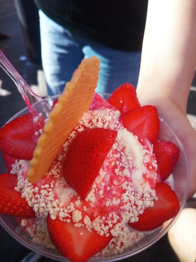 In a close-up, eye-level shot, a clear plastic cup filled with strawberry ice cream, fresh strawberries, and white crunchy toppings is held by a person. A light pink plastic spoon is partially visible, inserted into the cup. The ice cream is topped with a red strawberry sauce and a triangular waffle crisp. Sliced strawberries are arranged around the perimeter of the cup, with some partially submerged in the ice cream. The background is out of focus, showing a person wearing blue jeans and a dark top, with an arm extended, supporting the cup. The scene appears to be outdoors, possibly in a park or street in the city of Bad Homburg vor der Höhe, Germany, given the natural light and casual attire of the person.