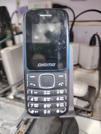 This is a medium shot, eye-level, close-up photograph of a black Digma feature phone, presented vertically on a dark gray stand. The phone has a blue trim around its screen and a metallic silver navigation button. The screen displays a reflection and appears smudged with dust. Below the screen, the numeric keypad is visible with white characters against black buttons. The phone is positioned in front of a blurred background that includes electronic components, a white boxy object, and cables. The overall scene suggests an indoor environment, possibly a repair shop or a cluttered workspace. There are no people or animals present in the image. The phone is the primary subject. The lighting is uneven, with some areas of the phone being brighter than others. The image is grainy and shows some motion blur. The location is identified as Bokhtar, Tajikistan.