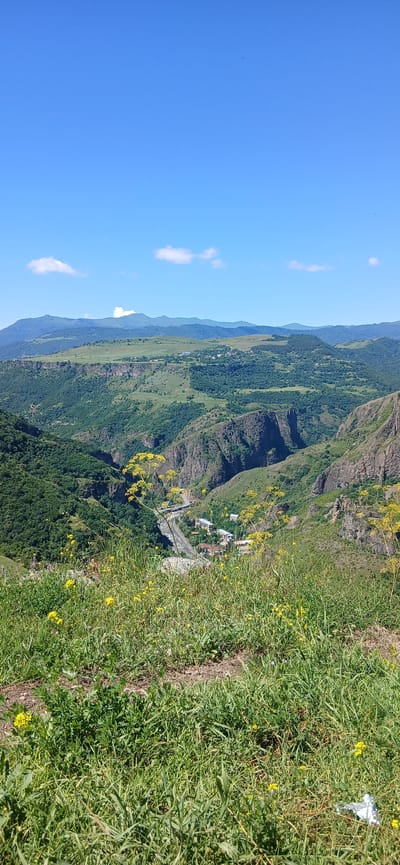 The image captures a panoramic view of a lush, verdant valley with rugged, steep cliffs on either side. In the foreground, tall green grass and yellow wildflowers blanket the terrain, suggesting a natural, undeveloped area. A winding road with a few scattered buildings is visible in the middle ground, nestled at the base of the valley, indicating a settlement. The background is dominated by rolling hills and distant mountains, fading into a clear blue sky with a few scattered white clouds. The scene is devoid of any human presence. The overall impression is one of natural beauty and tranquility. This landscape is characteristic of the region surrounding Dsegh, Armenia, a village known for its picturesque scenery and proximity to historical sites.