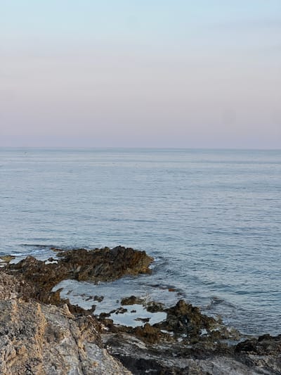 A scenic shot depicts a body of water meeting a rocky coastline. The water, appearing serene with faint ripples, stretches to the horizon under a pale sky. On the right, rugged rocks form the coast, their texture a mix of greys and browns with patches of white. The composition is such that the sky and water dominate the left, while the rocks occupy the right.