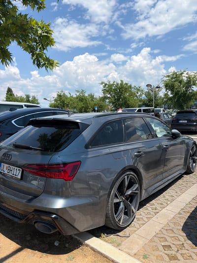 A dark gray Audi RS 6 Avant is parked on a cobblestone surface in a parking lot. The car is viewed from its rear quarter, with the rear passenger side door and the tailgate clearly visible. The license plate reads "UL BH117". In the background, other vehicles, including a black Porsche Cayenne with license plate "UL BJ846", are parked. Trees and a bright blue sky with scattered white clouds form the backdrop. The scene appears to be in Mojanovići, Montenegro.
