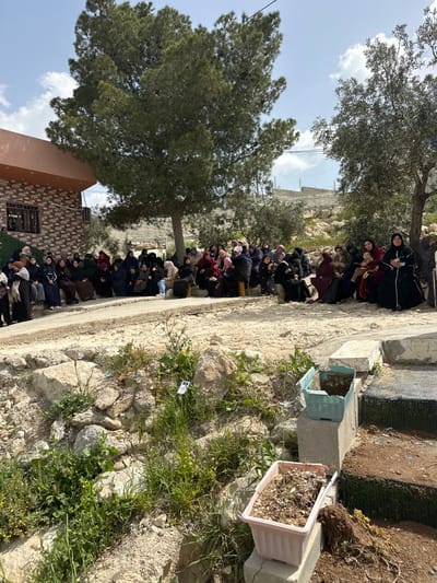 The image depicts an outdoor gathering of approximately 50-70 individuals, predominantly women and children, in the city of Dura, Palestinian Territory, under a partly cloudy sky.
In the foreground, an uneven patch of ground shows rocks, sparse soil, and various green plants and weeds. Two plastic planters, one light green and one light pink, rest on concrete blocks on the right side of this area, containing soil and minimal plant growth.
The midground features a large group of people seated on a slightly elevated, light-colored dirt or paved surface. Most individuals are women, wearing traditional attire including headscarves and long garments, primarily in dark colors such as black, brown, and navy, with some accents of red or lighter shades. Several children are present, some held by adults. The group appears to be observing or listening, with their attention generally directed towards the left side of the frame.
The background includes a two-story building on the left with a patterned brick-like facade and a light orange roof overhang, featuring an externally mounted air conditioning unit. A large, dense evergreen tree stands centrally, providing shade over a portion of the gathering. To the right, another olive tree is visible. Beyond the gathering and trees, additional residential structures and rocky, uncultivated hills extend towards the horizon. Utility lines are visible traversing the upper portion of the sky.