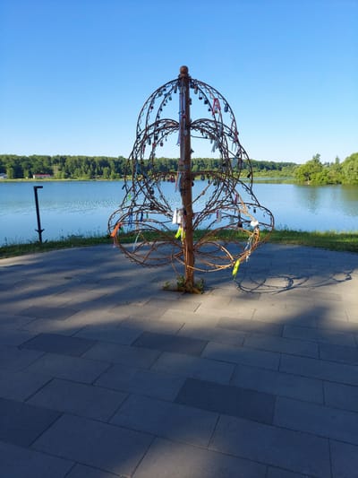 A rustic, metal sculpture, resembling a stylized tree adorned with numerous love locks and colorful ribbons, stands on a paved walkway beside a tranquil lake. The sculpture is made of intertwined metal rods, with an opening at its center, and is anchored in the ground with some vegetation growing around its base. In the background, a dense forest line borders the far side of the lake, under a clear, bright blue sky. The paved area exhibits a pattern of rectangular stone tiles, with dappled sunlight casting shadows. A dark pole-like object, possibly a light fixture or signpost, is visible to the left of the sculpture, near the water's edge. The overall scene suggests a peaceful outdoor location, likely in the city of Razvetye, Russia, commonly used as a scenic spot for couples.