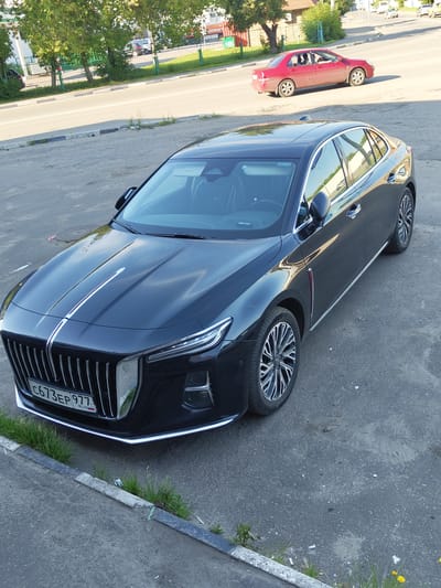 A black sedan is parked on an asphalt road in Zhukovsky, Russia. The car is facing right, and its front grille, headlights, and license plate are visible. The license plate reads "C673EP 977 RUS". A red sedan is driving on the road in the background, also facing right. Trees and buildings are visible in the background. The sun is shining, casting shadows on the ground.