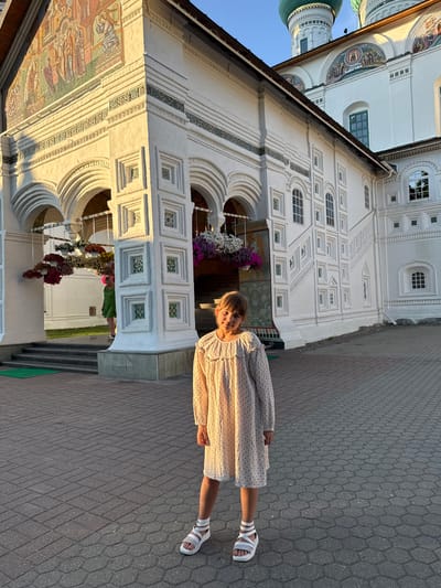 The image displays a young girl, approximately 7-10 years old, standing in the foreground, facing the camera with a slight smile. She has light brown hair with bangs and is dressed in a long-sleeved, cream-colored dress featuring small dark polka dots and ruffled details around the neckline and cuffs. She wears white platform sandals over white socks.
Behind the girl is a large, multi-story white building, characterized by intricate architectural details including arches, pilasters, and decorative square patterns. The building exhibits multiple colorful mosaic murals; a prominent, multi-figure mosaic is on the upper left section of the facade, and a smaller mosaic is visible on a higher structure. Two green onion domes rise above the white walls against a clear blue sky on the upper right. Several hanging flower baskets containing white, pink, and purple blooms are suspended near arched entrances on the left side of the building. Another person, visible from the back and side in a green dress, is ascending a set of steps into one of these arched doorways on the left.
The ground in the foreground is paved with grey hexagonal interlocking pavers. The scene is illuminated by warm, low-angle sunlight, indicating late afternoon or early evening. This building is located in Kuznechikha, Russia.