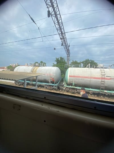 This image, taken from inside a train, shows a view of the exterior scenery. The primary focus is on a train with several large, cylindrical tank cars. These cars are light gray with red and green markings. Visible text on one of the cars includes "76605369" and other Cyrillic characters. A man with a beard, wearing a dark cap, is visible near one of the tank cars.
In the background, there are trees and buildings. Above the train, a complex network of overhead power lines and a lattice tower are prominent, suggesting the train is on an electrified railway line. The sky is overcast with clouds. The image is taken during the daytime. The overall scene depicts a railway yard or station with trains parked on tracks.