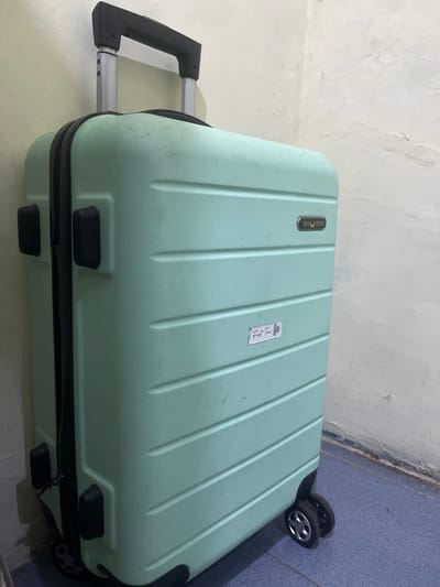 A mint green suitcase stands upright against a textured, off-white wall. The suitcase has a patterned surface with horizontal ridges and two black handles on the side. A silver telescoping handle extends from the top. Two small black protrusions are visible on the left side of the suitcase, likely for carrying. A rectangular black logo with gold lettering reading "POLO VIENNA" is affixed to the upper right quadrant of the suitcase. Below this logo, a small white sticker with handwritten text that reads "First Time" and a flower design is attached. The suitcase rests on four black wheels at the bottom, two of which are clearly visible. The floor is a blue speckled surface. The wall shows signs of wear, with some discoloration and marks. The scene is indoors, likely a room, with the lighting casting shadows on the wall. There are no people or animals present in the image. The image is set in Anggaswangi, Indonesia.