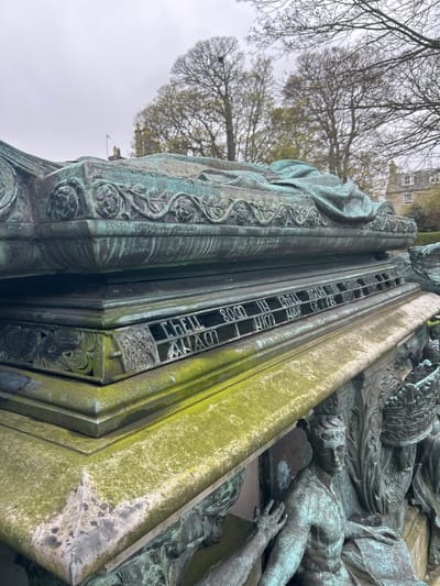 This image shows a section of a weathered, green-tinged bronze monument. The inscription "CHELL 8000 IN CHELL AGCH IN DHONS" is clearly visible, along with other indecipherable phrases due to wear and the angle of the photo. To the right of the inscription, there are sculptural representations of people. The overall environment suggests an outdoor location, likely within the city of Aberdeen City, United Kingdom.