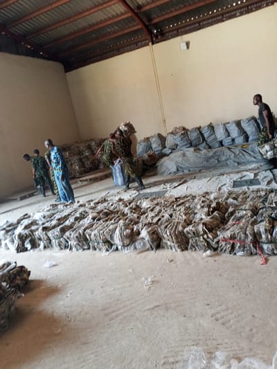 The scene depicts a large indoor space, possibly a warehouse, in Pasere, Nigeria. Five individuals are present: four appear to be soldiers in military fatigues, and one civilian wearing a blue patterned shirt and jeans. The soldiers are handling and moving bundles. The civilian is standing observing. The warehouse floor is covered with rows of tightly packed bundles, possibly containing agricultural products, wrapped in plastic and tied with string. Additional bundles are stacked towards the back of the warehouse and covered with a dark-colored tarp. The warehouse has a corrugated metal roof and light-colored walls.
