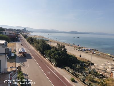 An aerial, high-angle medium shot captures a coastal scene in Follonica, Italy, on a bright, clear day. The image features a wide sandy beach with numerous beach umbrellas and lounge chairs, many of which are unoccupied. The gentle blue sea stretches out to the horizon, meeting a hazy mountain range in the distance. A faint contrail from an aircraft is visible in the pale blue sky.

In the foreground, a paved pedestrian and bicycle path runs parallel to the beach, separated by a strip of trees and shrubs. The path is marked with white lines and bicycle lane indicators. A person is seen riding a bicycle on the path. Adjacent to the path, a small white tent is set up with tables and chairs underneath, possibly a food vendor. Two bicycles are parked near the tent.

Further back, a street is visible with buildings lining one side. These buildings appear to be a mix of residential and commercial structures, with some taller buildings in the background, forming part of the city's skyline. The overall impression is of a serene, well-maintained beach resort area.