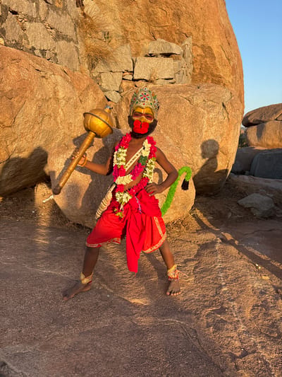 A young boy in a costume resembling the Hindu god Hanuman poses on a rocky terrain. He is dressed in red clothing, adorned with floral garlands and an ornamental headdress. His face is painted with yellow makeup and red around the mouth. He holds a golden mace in his left hand. A green and black faux tail is attached to his back. The scene is set against a backdrop of large reddish-brown boulders. The image was taken outdoors, with natural lighting. The location appears to be in the city of Kamalapura, India, given the characteristic rock formations.