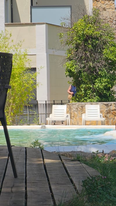 The image depicts an outdoor scene, likely a patio or poolside area, at Alhama De Aragón, Spain. A person, seen from the back, is leaning over and appears to be holding a blue bag and a straw hat or basket. They are positioned near a lush green tree with a stone wall in the background. To the right of the person, white plastic lounge chairs are set up next to a pool with a tiled mosaic bottom. The water in the pool has a bright turquoise hue. A wooden walkway made of planks runs along the edge of the pool, with grass and small plants growing in the gaps and along the sides. The scene is bathed in sunlight, suggesting it is daytime. The overall impression is one of relaxation and leisure in a warm climate.