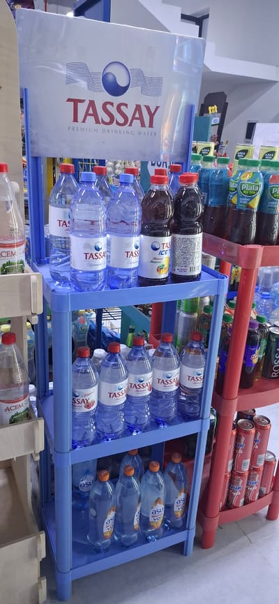 The image shows a retail display of bottled beverages inside a store. The main focus is on a blue plastic shelving unit filled with various bottled drinks. On the top shelf, there are several large bottles of "TASSAY" premium drinking water. The middle shelf displays more "TASSAY" bottles, including some with red caps and labels indicating flavors like "Малина" (Raspberry) and "Vitamin C". There are also two bottles of "TASSAY ICE" tea with mango and pineapple flavors. The bottom shelf of the blue unit contains more bottled drinks, some with orange caps and labels featuring a stylized sun graphic and text that appears to be "asu". To the right of the blue shelf, a red plastic shelf unit is visible, stacked with cans of "Fresh" and other unidentifiable beverages, as well as some bottles. In the background, other shelves with drinks are partially visible, along with store signage, including a large white banner with the "TASSAY" logo and text. The lighting suggests an indoor retail environment. The scene is set in the city of Arys, Kazakhstan, likely within a grocery store or supermarket. There are no people visible in the image.