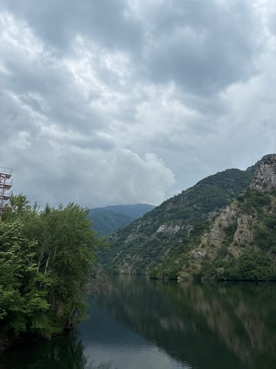 A vast, dark green lake stretches across the foreground, reflecting the overcast sky and the steep, tree-covered mountains that rise on either side. The mountains are rugged and rocky in places, with dense foliage clinging to their slopes. In the upper left portion of the image, a tall, metallic scaffolding structure is visible, partially obscured by lush green trees that overhang the water. The sky is a mottled grey, with large clouds creating a sense of depth and shadow. The overall scene depicts a natural landscape, likely a reservoir or a deep river, with man-made structure juxtaposed against the wild environment. This location is part of the Krichim, Bulgaria area.