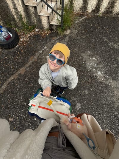 An overhead shot captures a young child smiling up at the camera, situated outdoors in Nerin, Montenegro. The scene is dominated by dark asphalt ground, showing patches of white residue and scattered dry leaves. To the left, concrete steps with a black metal railing ascend, bordered by sparse green grass.
The primary subject is a fair-haired boy, appearing to be a toddler or preschooler. He wears a bright yellow beanie, stylish blue-rimmed sunglasses, a light grey zippered jacket, and dark sneakers. He is holding a black and white electronic-looking device in his right hand. The child's gaze is directed upwards towards the camera, and he has a clear, happy smile on his face.
Partially visible on the right side of the frame is an adult, likely holding the camera, indicated by a leg in grey trousers, an arm or body covered by a cream-colored fluffy garment, and a beige canvas bag with brown accents. Attached to this bag are a blue plastic clip and a small, light-haired doll with a flower on its head, resembling a Troll doll.
Next to the child, a beige and colorful backpack or a part of a stroller with red zippers, a yellow strap, and blue and green sections is visible. In the lower left corner, an old black car tire lies on its side, serving as an impromptu container for several plastic water bottles, some clear and some red.
The time of day appears to be daytime, with diffused light suggesting either an overcast sky or indirect sunlight. No discernible text is visible on any objects. The overall impression is a casual, everyday outdoor moment captured from a unique perspective.
