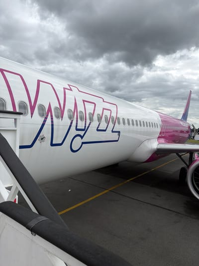 A Wizz Air airplane is depicted parked on an airport tarmac under a heavily overcast, grey sky. The image is taken from a low-angle perspective, looking up and along the side of the aircraft's fuselage. The plane's livery is primarily white, adorned with a large, stylized "Wizz" logo outlined in magenta and blue on the fuselage. The rear section of the fuselage and the vertical stabilizer (tail fin) are solid magenta, with "Wizz Air" text visible on the tail.

Numerous passenger windows are visible along the length of the fuselage. In the upper right of the frame, a portion of one of the aircraft's engine nacelles can be seen. The ground beneath the plane is dark grey tarmac, marked with yellow lines. A structure resembling boarding stairs or a jet bridge is present in the foreground, connecting to the aircraft, suggesting either passenger boarding or disembarkation is underway or imminent. A small circular gauge or port is visible on the fuselage near the blue part of the "Wizz" logo.

The weather appears to be a cloudy day, with no direct sunlight. No people are explicitly visible. The scene captures an industrial aviation environment, likely at an airport in Tillé, France.