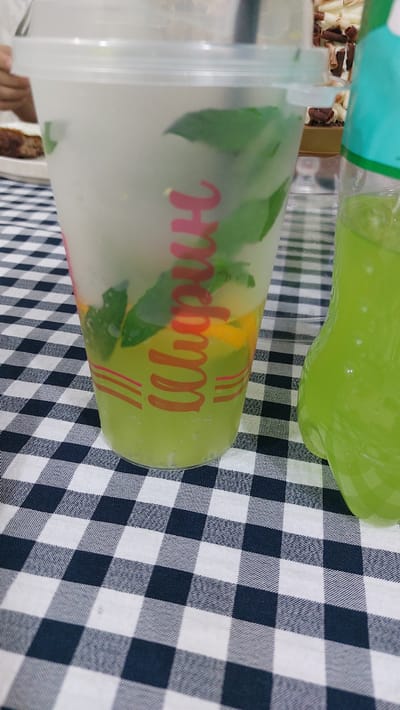 A close-up shot shows a clear plastic cup filled with a green beverage and mint leaves. The cup has a pinkish-red logo with swirling letters. Next to it, on the right, is a green bottle also filled with a similar green liquid. Both items are placed on a black and white checkered tablecloth. In the background, a blurry image of what appears to be food and a dessert can be seen. The scene is likely in a restaurant or cafe setting in the city of Kara-Balta, Kyrgyzstan, given the context provided.