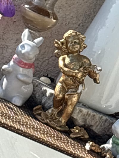 This close-up, slightly overhead shot features miniature decorative figures on a textured, neutral-colored surface, likely a shelf or display. In the foreground and center, a small, gray ceramic bunny figure with large ears and a red ribbon around its neck faces to the left. To the left of the bunny, a shiny, brown, bulbous object, possibly a miniature vase or part of another decorative piece, is visible.

Dominating the upper portion of the image is a golden, metallic figurine of a winged cherub or angelic figure in a reclining position, its body positioned diagonally across the frame. The cherub appears to be interacting with another small, dark-colored object, possibly a part of the same figurine or another element in the display. The cherub's metallic surface is worn, showing hints of its original polished sheen and some lighter, possibly white, paint or material.

Behind the cherub, a rough, gray, rock-like texture suggests a landscape or background element. To the right of the cherub, a textured surface with a pattern resembling bark or small, raised dots in shades of brown and gold adds another layer to the display. The lighting is bright, casting subtle shadows and highlighting the metallic sheen of the cherub and the textured background. The overall impression is of a collection of small, decorative collectibles, possibly part of a larger display or diorama. No text is visible in the image. The location context of Osterode am Harz, Germany, does not offer specific visual cues within the image itself.
