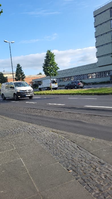 The image shows a street scene in Schweinfurt, Germany, at the coordinates 50.036, 10.211. A white van is driving on the road, headed towards the viewer. A second, white van is parked on the right side of the road, with a black car parked behind it. A tall building is visible in the background, on the right side of the image. The building has a modern design, with a light gray facade. There is a single tree in the foreground, and a light pole with a streetlight. The street has a cobblestone sidewalk on the left and a paved shoulder on the right. The sky is blue, with a few white clouds.