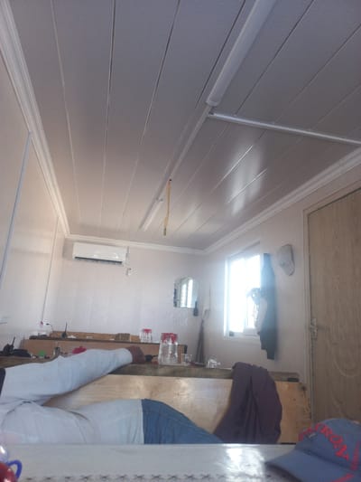 The image shows the interior of a room, possibly a temporary structure or a utilitarian space. The ceiling is paneled with white material, and a fluorescent light fixture runs parallel to the panels. An air conditioning unit is mounted high on a light-colored wall. To the right, a wooden door with a simple design is visible. A window with natural light streaming through is also present on the right side of the room. In the foreground, a person is seated, wearing a white long-sleeved shirt and blue jeans. Their arm rests on a wooden surface, which appears to be part of a bench or table. On this surface, several objects are scattered, including clear glasses, a red container, and some electronic devices. A dark-colored garment is draped over the edge of the bench. A blue baseball cap with red lettering is also visible in the lower right corner. The overall impression is of a modest, functional living or working space. The location is stated to be in the city of Şəfalı, Azerbaijan.