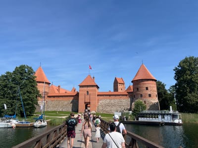 The image captures a view of Trakai Island Castle in Trakai, Lithuania, on a clear, sunny day. A wooden bridge, teeming with people walking towards the castle, spans a body of water. The castle itself, constructed of red brick and stone, features multiple towers with conical, orange-tiled roofs. A red flag with a white emblem flies from one of the towers. On the water to the left, several sailboats are docked, and to the right, a white tour boat named "AKMENA" is moored. Lush green trees surround the castle and line the water's edge.
