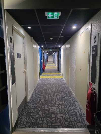 An elongated hotel corridor receding into the distance. On the left, dark blue paneled walls with emergency exit signs and inset lighting. White doors with room numbers line both sides of the hallway. The carpet is a pattern of symbols and letters in grey. A yellow stripe runs down the middle of the corridor. The perspective is slightly rotated, creating a warped effect, with a focus on the receding vanishing point.