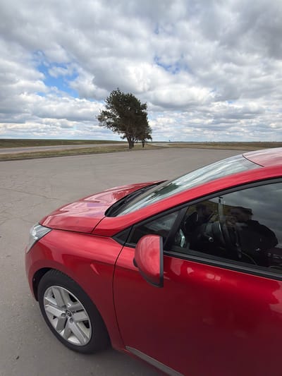 A close-up, angled shot captures a red car parked on an expansive paved surface, possibly an airfield or large parking area. The sky is heavily clouded with patches of blue visible. A single, mature tree stands at a distance across the tarmac. The environment appears to be in a rural location potentially within the Абухаўскі сельскі Савет area of Belarus.