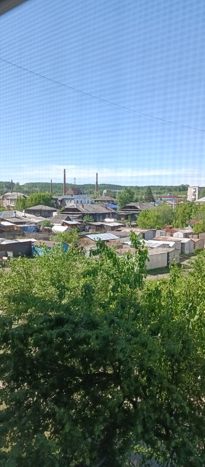 The image shows a view of a small urban area in Irbit, Russia, characterized by a mix of residential and industrial elements. In the foreground are the tops of trees. The middle ground reveals a dense arrangement of single-story buildings, including homes with weathered wooden structures and corrugated metal roofs, as well as some small garages or sheds. In the background, several brick industrial chimneys rise above the treeline, indicative of some kind of factory or plant. The sky is clear and blue. The buildings suggest a lower-income residential area.