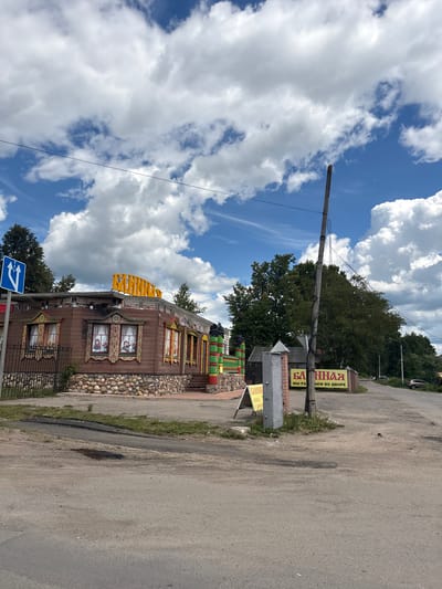 The image captures a scene in Pereslavl-Zalessky, Russia, on a bright, partly cloudy day. The sky is a vibrant blue with large, fluffy white clouds. A wooden pole with wires stretches across the frame, suggesting the presence of electricity or communication lines.
In the foreground, a distinctive wooden building stands out. It is adorned with colorful, ornate window frames and a stone foundation. The building's architecture has a traditional Russian feel, with intricate carvings and decorative elements. A bright yellow structure, possibly a sign or part of the building's facade, is visible on the roof. A black metal fence runs along the front of the building.
To the right of the building, a paved road is visible, with patches of grass and some minor damage to the asphalt. There are also several signs with Russian text. One prominent yellow sign reads "БАННАЯ" (BATHHOUSE) with "ЕМ ВО ДВОРЕ" (IN THE YARD) below it. Another sign, partially visible, seems to have "МЫ РА" on it. Further down the road, a traffic sign with blue arrows indicates a direction. Trees and foliage line the background, and a car can be seen in the distance on the road. The overall impression is a sunny day in a semi-rural or suburban setting, with a focus on a unique, decorated building.