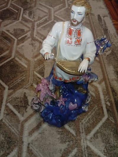 A porcelain figurine of a man playing a stringed instrument stands on a patterned rug. The figurine depicts a man with blonde hair, a beard, and blue eyes, wearing a white shirt with red embroidery and a blue skirt. He is holding a golden stringed instrument. The base of the figurine is adorned with blue and purple accents and floral elements. The rug has a geometric pattern in shades of brown and beige. The background is out of focus, showing a wooden floor. No specific location in Iskitim, Russia, can be determined from the image.