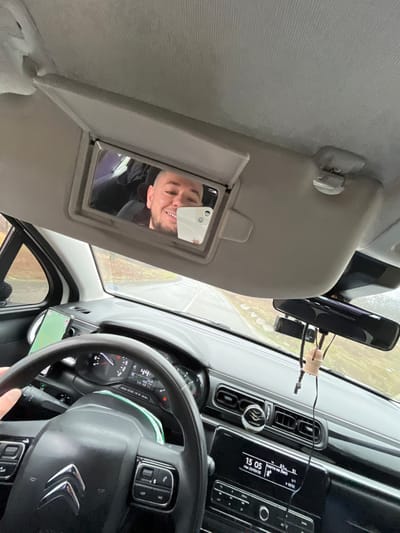 The image depicts the interior of a vehicle from the driver's perspective, capturing a male individual taking a selfie in the sun visor mirror while driving in Sredna mahala, Bulgaria.
A male individual with a shaved head and a beard is reflected in the overhead sun visor's vanity mirror. He is smiling and holds a white smartphone (visible by its multiple camera lenses) in his right hand, positioned to capture a selfie of himself. The driver's left hand is visible gripping the left side of the black multi-functional steering wheel, which features a Citroën logo.
The dashboard includes an instrument cluster with a digital display showing "44" (likely speed). A second smartphone, displaying a map application, is secured in a mount on the dashboard to the left of the steering wheel. The center console houses an infotainment system displaying the time "15 05" and date "Mon 23/03," along with Cyrillic text "РЕДНО НА ЛИС" and "02:52." A rearview mirror is present, with a black dashcam attached beneath it and a wooden bead air freshener suspended from it. The vehicle's headliner and sun visor are grey.
Through the windshield, a two-lane asphalt road is visible, appearing wet and curving to the right. The road is bordered by a brown, sparse roadside verge leading to bare-branched trees and shrubs under an overcast, light grey sky. The environment suggests a rural or semi-rural setting.