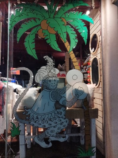 A storefront window displays a painted scene of a monkey in costume playing cymbals under a palm tree. The monkey wears a hat with a feather and a tunic with decorative trim. The palm tree has green fronds and brown coconuts. Behind the monkey, there's a white bench and some grass. Inside the store, clothing and home decor items are visible. Two oval mirrors hang on the wall to the right of the display.