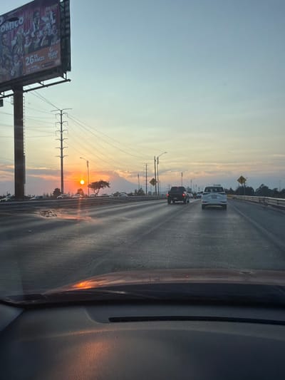 A photo taken from inside a car shows a multi-lane highway at sunset. Cars are in the distance. Power lines and streetlights run alongside the road. A billboard is visible on the left. The sun is a bright orange orb. The sky is a gradient of blue and orange. The dashboard of the car is visible in the foreground. The image is slightly blurry.