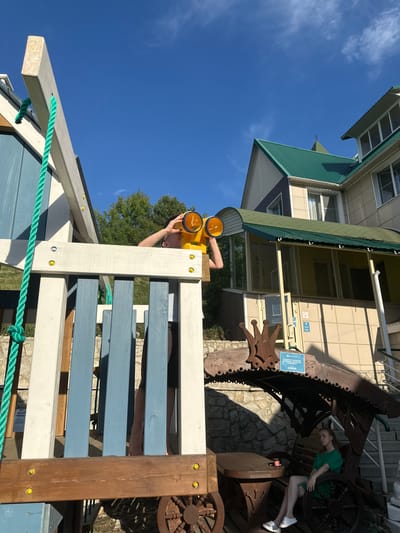 In the city of Danilovka (Altai), Russia, a young person, wearing a white t-shirt and dark shorts, stands on a wooden playground structure, looking through yellow binoculars. The playground features blue and white painted wooden planks and a green rope. In the background, a multi-story building with a green roof and beige siding is visible, along with trees and a clear blue sky. To the right of the playground, a young woman wearing a green dress sits on a wooden bench under a decorative wooden canopy. A stone wall and stairs are also present in the background.