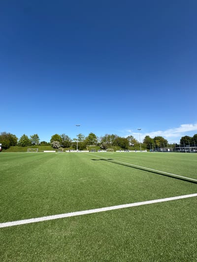 The image shows a football field in Wassenberg, Germany, on a clear, sunny day. The artificial green turf is marked with white lines. Several goals line the edge of the field. Light poles are visible in the background, along with trees. The sky is blue with a few scattered clouds. No people are visible in the frame.