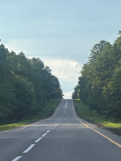 The image captures an asphalt road winding through a dense forest. The road is marked with a broken white line down the center and solid yellow lines along its edges. Lush green trees line both sides of the road, creating a tunnel-like effect as the road curves and ascends into the distance. The sky above is a pale blue with scattered white clouds, suggesting a temperate daytime. The scene appears to be a rural road within the Акцябрскі сельскі Савет, Belarus, given the characteristic landscape of Belarusian countryside. There are no people or vehicles visible in the image, and the environment is natural and unpopulated.