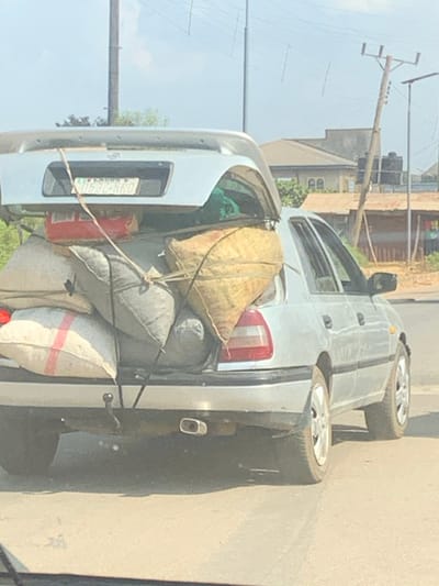 A silver sedan, likely a Nissan model, is depicted from a rear-quarter perspective on a bright day in Egbedi, Nigeria. The vehicle is stationary or moving slowly on an asphalt surface, heavily overloaded in the trunk area. The car's original trunk lid is detached and positioned horizontally on top of the stacked cargo, displaying a Nigerian license plate "LAGOS 2 D532 KRO." The open trunk is filled with numerous large, irregularly shaped sacks and bags; colors include light grey, beige (one with visible text), and white with a red stripe. A red rectangular container and green fabric are also visible amidst the load. Manila-colored ropes secure the cargo and the detached trunk lid to the vehicle. The car's rear features a single exhaust pipe and a tow hitch. In the background, under a clear sky, are utility poles with overhead power lines, residential-style buildings with light-colored roofs, and a smaller structure with a corrugated metal roof and visible black water tanks. No individuals are discernible within the frame.