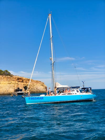 A bright blue catamaran named "FINISMAR" sails on the choppy, blue waters of the Atlantic Ocean, near the coast of Lagoa, Portugal. The boat is filled with passengers, many of whom are dressed in swimwear and appear to be enjoying a sunny day trip. A large, tan-colored cliff face, typical of the Algarve region, rises from the water to the left of the catamaran, dotted with green vegetation. Another boat, a dark gray catamaran, is anchored near the cliffs, and several people are seen kayaking in the water between the two vessels. The sky above is a clear, vibrant blue with a few wispy clouds. The overall scene depicts a recreational outing on the coast.