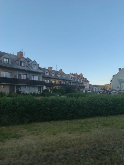 The image shows a row of attached residential buildings with red-tiled roofs and dormer windows. The buildings are painted in muted colors such as beige, gray, and yellow, and they have dark-colored window frames. A dense green hedge runs along the front of the buildings, and behind it, there is a patch of dry grass. In the background, there is a street with parked cars and more buildings, some of which are taller and have lighter facades. The sky is a clear, pale blue. The scene appears to be a street in a residential area of Chvalšiny, Czechia, possibly in the late afternoon or early evening.