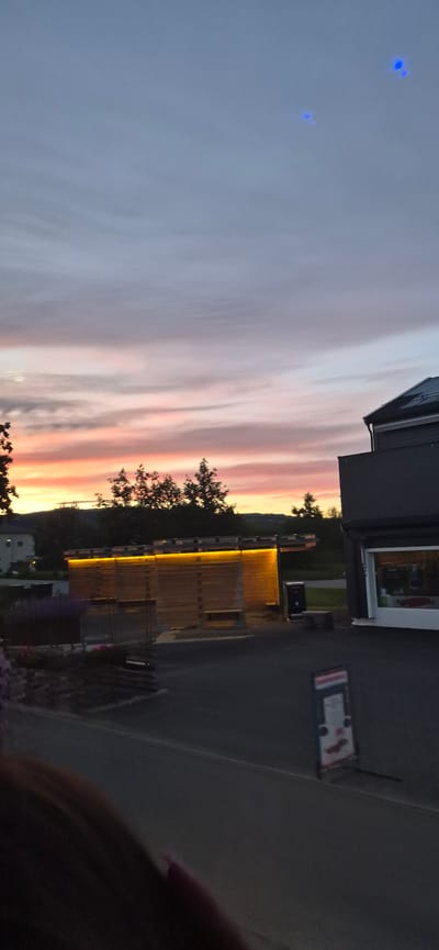 The image depicts a twilight scene in Biri, Norway, with a striking sky painted in hues of pink, orange, and purple, indicating the sunset. A modern building with a wooden facade and illuminated by warm yellow lights is prominently featured. The building appears to be a commercial establishment, possibly a shop or restaurant, with a large window visible. In the foreground, to the right, a person's shoulder and hand are visible, suggesting they are taking the photograph. The ground in front of the building is paved, and there are hints of greenery and trees surrounding the structure. The overall atmosphere is serene and beautiful, capturing the magical light of dusk. There is a sign on the wall of the building that is not legible. Two small blue lights are visible in the sky.