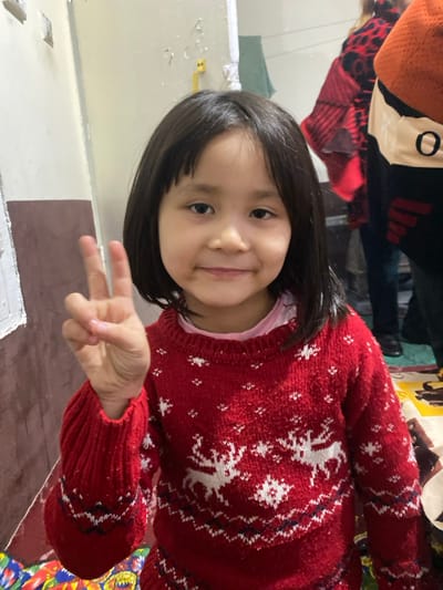 An image captured in Akhtachi, Uzbekistan, depicts a young girl posing indoors. The girl, with dark, straight, shoulder-length hair and bangs, is centered in the frame, facing forward with a slight smile. She is wearing a red knitted sweater adorned with white reindeer and snowflake patterns, over a light pink undershirt. Her right hand is raised, forming a V-sign with extended index and middle fingers.
The background consists of light-colored walls, displaying minor imperfections and a darker reddish-brown panel along the lower left. A white frame or trim is visible on the left wall. In the blurred background to the right, partial views of two individuals are discernible. One person wears a dark garment with a tan section featuring a black circular 'O' symbol. Another individual is seen wearing a red and black patterned garment. The bottom right shows a portion of a colorful, patterned floor covering.