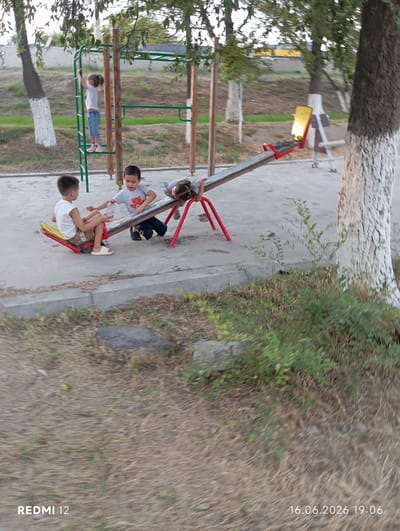In Bekabad, Uzbekistan, a scene unfolds at a playground with a seesaw as the central element. Three children are interacting with the seesaw. On the left, a young boy wearing a white t-shirt and brown shorts sits on one end of the seesaw, his feet resting on the ground. He holds a clear plastic bottle. To his right, another boy, also in a white t-shirt adorned with a red bear graphic and dark pants, is seated on the seesaw. He appears to be reaching towards the boy with the bottle. On the opposite end of the seesaw, a third child, partially visible, is lying down, with their head towards the seesaw's support. In the background, a young girl in a white t-shirt and blue jeans is climbing a set of monkey bars. The playground surface is a light-colored concrete, with patches of dry grass and green bushes surrounding the equipment. A large tree with a white-painted trunk stands to the right of the frame. The image is dated 16.06.2026 at 19:06.