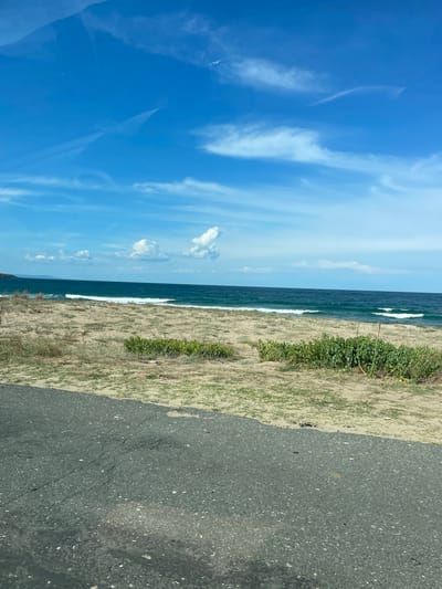 The image shows a view of the Black Sea coast near Dyuni, Bulgaria. The scene is captured from a slightly elevated perspective, likely from a road. In the foreground, there is a cracked asphalt road, partially visible. Beyond the road, a sandy beach stretches towards the sea, dotted with sparse, dry grass and patches of green vegetation. The sea is a deep blue-green, with gentle waves breaking on the shore, creating white foam. The horizon is marked by the meeting of the sea and the sky, with a distant coastline visible on the left. The sky is a vibrant blue, with scattered white clouds, some of which are wispy and thin, while others are more cumulus-like. The overall impression is a serene coastal landscape under a bright sky.