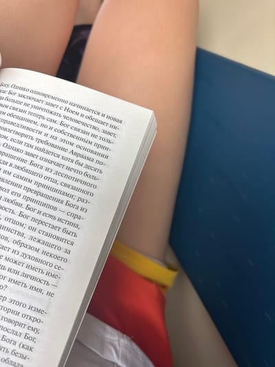 An overhead view captures the upper thighs of a person holding an open book. The book, with white pages and a visible spine on the left, displays Russian text on its right-hand page, discussing theological themes such as "God," "covenant," "Noah," and "Abraham." The person's bare skin is visible on the thighs, with a dark garment appearing on the upper left thigh and a red garment featuring a yellow band on the upper right thigh. A dark blue surface is present to the right of the book, with a lighter, beige-toned area above it, forming the background. The location in the city of Biryusinsk, Russia, is not discernible from the visual evidence within the image.