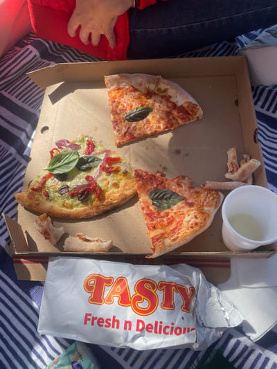The image captures a close-up, overhead view of a pizza box resting on a blue and white striped blanket, likely in an outdoor setting. Three slices of pizza are visible within the open cardboard box. One slice, in the upper right, is a classic margherita with cheese and tomato sauce, garnished with a single basil leaf. Another slice, to the left, is topped with pesto, sun-dried tomatoes, red onion, olives, and basil. A third slice, at the bottom right, is similar to the margherita but appears to have slightly more cheese and sauce. Several small, crusty remnants of pizza are scattered in the box. To the right of the pizza, a small white plastic cup contains a pale yellow liquid, possibly a drink. In front of the pizza box is a white paper bag with the word "TASTY" in large red and yellow lettering, along with the text "Fresh n Delicious". A hand, partially visible in the upper left corner, appears to be resting on a red fabric item, potentially a jacket or blanket. The lighting suggests it is daytime, with shadows cast by the objects. The context of the city of Gold Coast, Australia, is not explicitly depicted in the visual elements of the image itself.