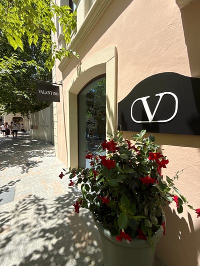 An outdoor shot captures the facade of a Valentino store in La Roca del Vallès, Spain. A black sign with "VALENTINO" in white lettering hangs from an ornate metal bracket. To the right, a large black sign features a white "V" logo. In the foreground, a potted plant with vibrant red flowers and green leaves is positioned near the store entrance, casting a shadow on the light-colored textured wall of the building. The ground is paved with light-colored stones, and dappled sunlight filters through the leaves of trees overhead, creating a pattern of shadows on the walkway. Several people are walking along the sidewalk in the background, some carrying shopping bags. A temporary barrier with Valentino branding is visible along the sidewalk to the left.
